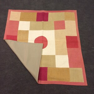 Może przedstawiać: Kwadratowy dywan w stylu patchwork z czerwonym kołem w środku. Dywan wykonany jest z tkanin w r&oacute;żnych kolorach, w tym czerwonym, brązowym i białym.
