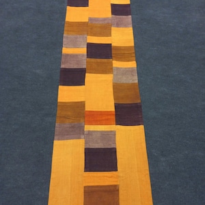 May include: A long, narrow patchwork runner with a geometric design. The runner is made of various shades of brown, orange, and purple fabric.