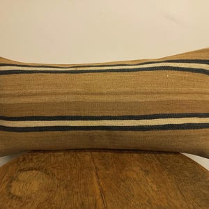 Peut inclure: Coussin rectangulaire à rayures horizontales dans les tons de beige, marron, noir et blanc. Le coussin est en tissu tissé et a un aspect texturé. Il est posé sur une surface en bois.