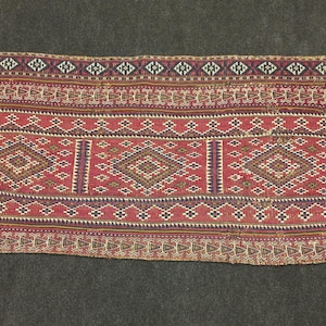 May include: A rectangular rug in shades of red, with detailed geometric patterns. The design includes repeating diamond shapes in brown and cream, framed by bands of smaller geometric patterns in multiple colours.