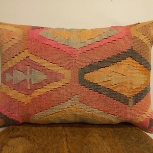 May include: Rectangular decorative pillow with a geometric pattern. The pillow features a woven design with a combination of pink, orange, gray, and brown hues. The pattern consists of diamond and chevron shapes, creating a textured appearance.