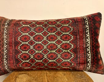 May include: Rectangular pillow with a geometric pattern in shades of red, brown, and cream. The pillow has a central design of diamond shapes and borders with vertical stripes and zigzags. The pillow is placed on a wooden surface.