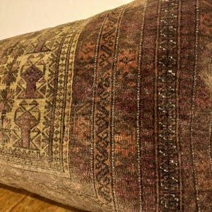 May include: A rectangular decorative pillow with a vintage, patterned design. The pillow features a mix of colors, including brown, beige, and burgundy. The design includes geometric shapes and vertical stripes, creating a textured appearance. The pillow is likely made of a soft fabric.