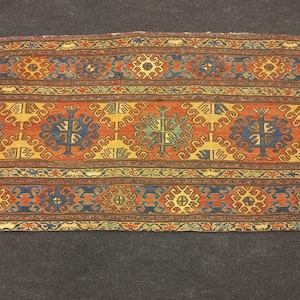 May include: Rectangular rug with a detailed, symmetrical pattern. The rug features a central field of orange with repeating blue and yellow tree-like motifs. Borders are in blue, yellow, and orange, creating a vibrant, traditional design.