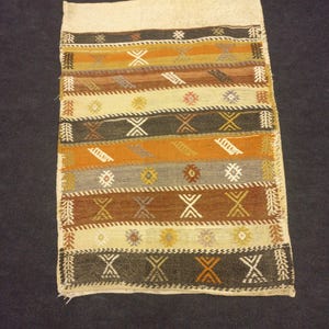 May include: A rectangular rug with a cream background and colorful geometric patterns in shades of brown, orange, gray, and black. The rug has a fringe border.