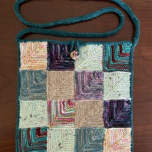Könnte beinhalten: Eine quadratische, gestrickte Umhängetasche mit einem Patchwork-Design aus sechzehn bunten Quadraten in den Farben Petrol, Beige, Lila und Creme. Die Tasche hat einen langen petrolfarbenen Riemen und einen einzelnen runden Knopfverschluss.