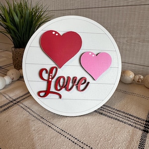 Peut inclure: Panneau rond en bois blanc avec un cœur rouge et un cœur rose. Le mot "Love" est écrit en script rouge. Le panneau est posé sur une surface à motifs beige et blanc.