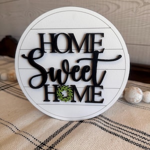 May include: Round wooden sign with the words "Home Sweet Home" in black lettering. The word "Home" is repeated and the second time it is decorated with a green wreath.