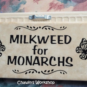 Op de afbeelding: Een rechthoekig bord met de tekst "MILKWEED for MONARCHS" in zwart. Twee zwarte vlindersilhouetten flankeren de tekst. Het bord heeft een cr&egrave;mekleurige achtergrond met een decoratieve rand en een metalen hanger.