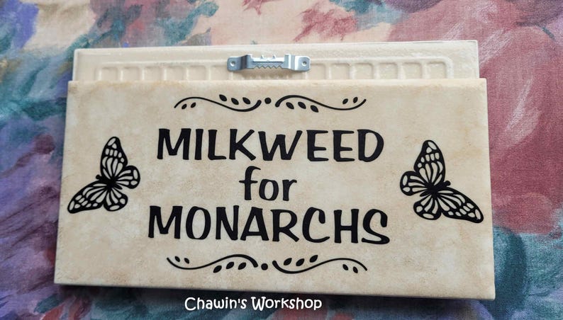 Op de afbeelding: Een rechthoekig bord met de tekst "MILKWEED for MONARCHS" in zwart. Twee zwarte vlindersilhouetten flankeren de tekst. Het bord heeft een cr&egrave;mekleurige achtergrond met een decoratieve rand en een metalen hanger.