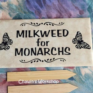Op de afbeelding: Een rechthoekige, cr&egrave;mekleurige tegel met de zwarte woorden "MILKWEED for MONARCHS", geflankeerd door vlindersilhouetten. Decoratieve krullen en twee houten plantenmarkeringen zijn ook zichtbaar.