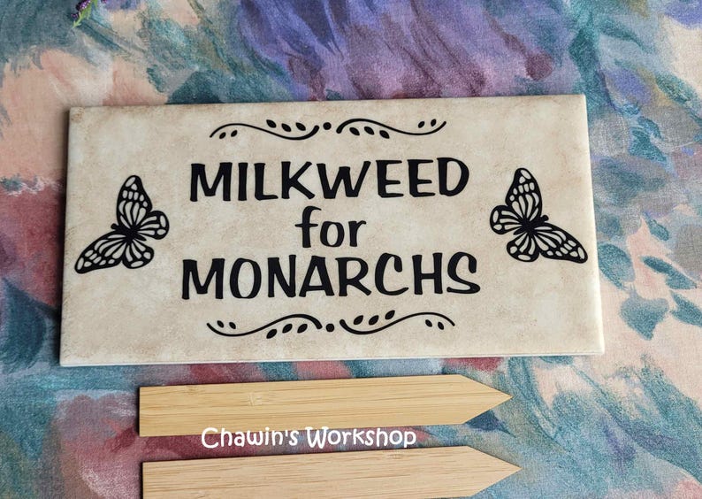 Op de afbeelding: Een rechthoekige, cr&egrave;mekleurige tegel met de zwarte woorden "MILKWEED for MONARCHS", geflankeerd door vlindersilhouetten. Decoratieve krullen en twee houten plantenmarkeringen zijn ook zichtbaar.