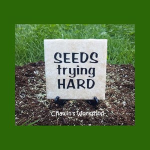 May include: A white square tile with black lettering that reads "SEEDS trying HARD" on a bed of dark brown soil with green grass in the background. The tile is on a black metal stand.