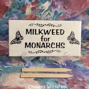 Op de afbeelding: Een cr&egrave;mekleurig rechthoekig bord met de tekst "MILKWEED for MONARCHS" in zwart, geflankeerd door vlinderillustraties. Het bord wordt weergegeven op een transparante standaard, met twee houten palen eronder. De tekst "Chawin's Workshop" staat onderaan.