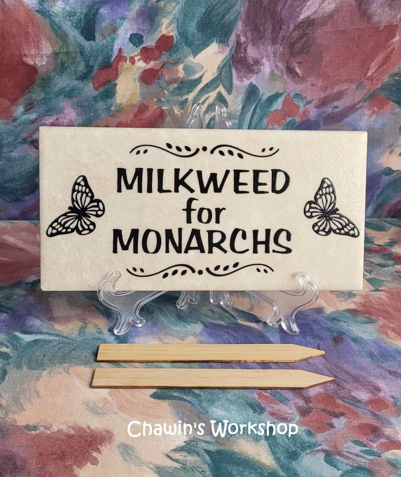 Op de afbeelding: Een cr&egrave;mekleurig rechthoekig bord met de tekst "MILKWEED for MONARCHS" in zwart, geflankeerd door vlinderillustraties. Het bord wordt weergegeven op een transparante standaard, met twee houten palen eronder. De tekst "Chawin's Workshop" staat onderaan.