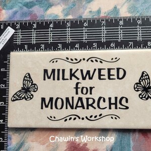 Op de afbeelding: Een cr&egrave;mekleurige rechthoekige tegel met de zwarte tekst "MILKWEED for MONARCHS" en vlinderontwerpen. De tegel is ongeveer 15 cm lang. De tekst "Chawin's Workshop" staat onderaan.