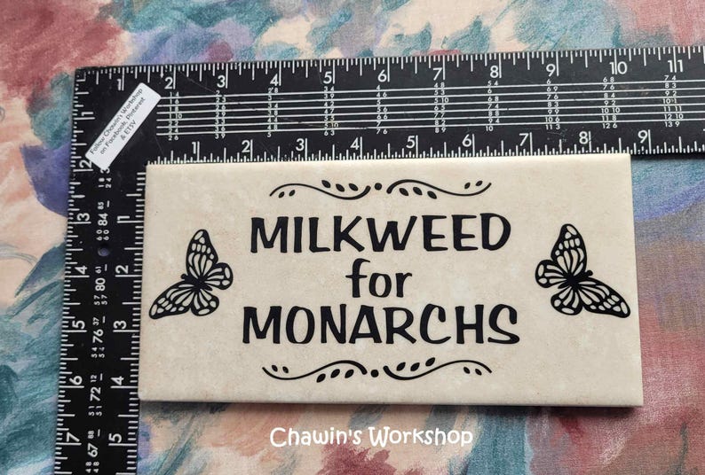 Op de afbeelding: Een cr&egrave;mekleurige rechthoekige tegel met de zwarte tekst "MILKWEED for MONARCHS" en vlinderontwerpen. De tegel is ongeveer 15 cm lang. De tekst "Chawin's Workshop" staat onderaan.