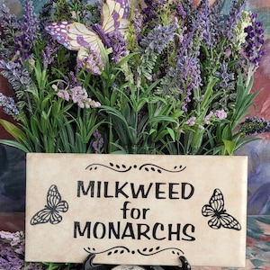 Op de afbeelding: Een rechthoekig bord met de tekst "MILKWEED for MONARCHS" en vlinderillustraties, omringd door paarse bloemen en groen. Een decoratieve vlinder en een zakhorloge zijn ook aanwezig.