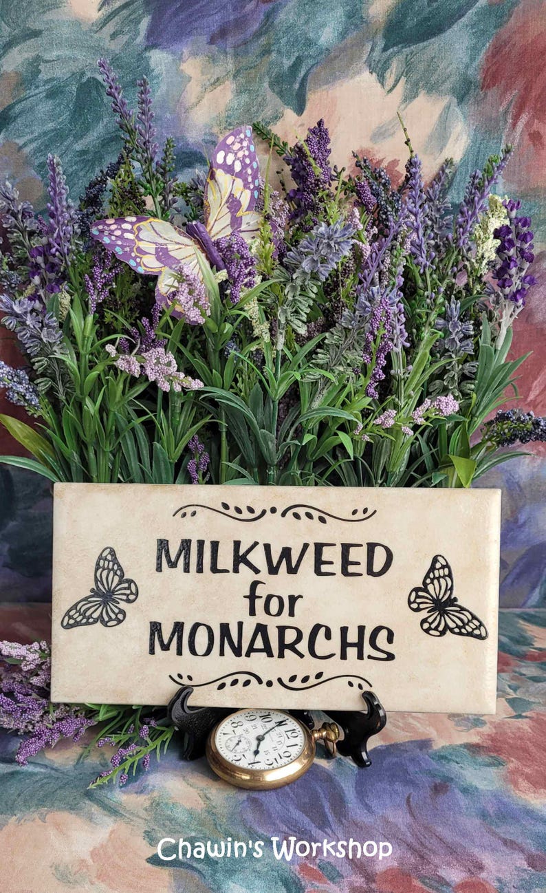 Op de afbeelding: Een rechthoekig bord met de tekst "MILKWEED for MONARCHS" en vlinderillustraties, omringd door paarse bloemen en groen. Een decoratieve vlinder en een zakhorloge zijn ook aanwezig.