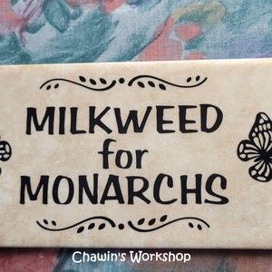 Op de afbeelding: Rechthoekig bord met de zwarte tekst "MILKWEED for MONARCHS", met vlinderillustraties. Het bord heeft een beige achtergrond met decoratieve krullen en de tekst "Chawin's Workshop" onderaan.