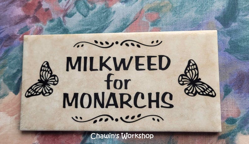 Op de afbeelding: Rechthoekig bord met de zwarte tekst "MILKWEED for MONARCHS", met vlinderillustraties. Het bord heeft een beige achtergrond met decoratieve krullen en de tekst "Chawin's Workshop" onderaan.