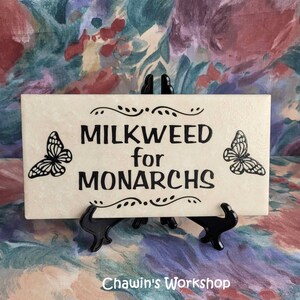 Op de afbeelding: Een rechthoekige cr&egrave;mekleurige tegel met de zwarte tekst "MILKWEED for MONARCHS". Twee zwarte vlinderillustraties flankeren de tekst. De tegel wordt gepresenteerd op een zwarte standaard. De tekst "Chawin's Workshop" staat onderaan.