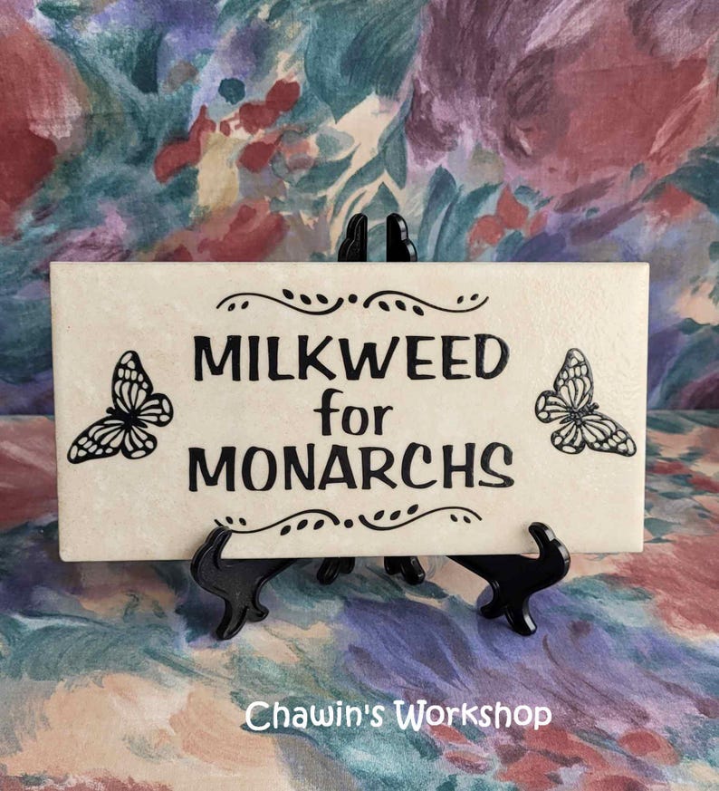 Op de afbeelding: Een rechthoekige cr&egrave;mekleurige tegel met de zwarte tekst "MILKWEED for MONARCHS". Twee zwarte vlinderillustraties flankeren de tekst. De tegel wordt gepresenteerd op een zwarte standaard. De tekst "Chawin's Workshop" staat onderaan.
