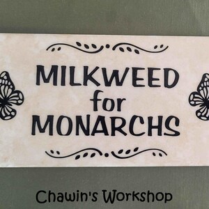 Op de afbeelding: Een rechthoekige, cr&egrave;mekleurige tegel met de zwarte tekst "MILKWEED for MONARCHS", geflankeerd door vlinderillustraties. Decoratieve versieringen en de tekst "Chawin's Workshop" zijn ook aanwezig.