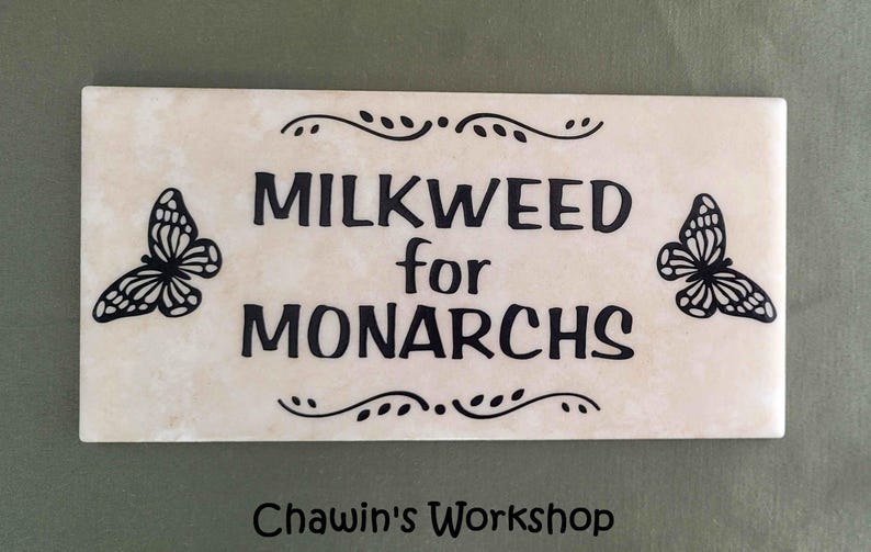 Op de afbeelding: Een rechthoekige, cr&egrave;mekleurige tegel met de zwarte tekst "MILKWEED for MONARCHS", geflankeerd door vlinderillustraties. Decoratieve versieringen en de tekst "Chawin's Workshop" zijn ook aanwezig.