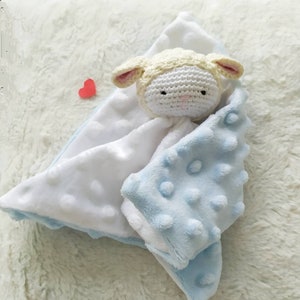 Op de afbeelding: Een babyknuffel met een gehaakt lammetjeshoofd. De deken is wit en lichtblauw, met een gestructureerd, gestippeld patroon op het blauwe gedeelte. Het lammetje heeft een cr&egrave;mekleurig hoofd en roze accenten op de oren.