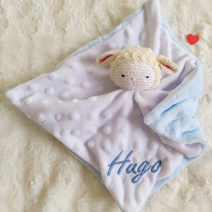 Op de afbeelding: Een witte en blauwe babydeken met een gehaakt lammetjeshoofdje. De deken heeft een zacht, getextureerd oppervlak, een blauwe rand en de naam "Hugo" geborduurd in blauw. Een klein rood hart is zichtbaar.