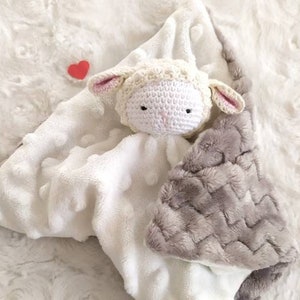 Op de afbeelding: Een witte en grijze babyknuffel met een gehaakt lammetjeshoofd. De deken heeft een zacht, getextureerd oppervlak met een grijs chevronpatroon op een hoek en een klein rood hart. Het lammetje heeft roze oren.