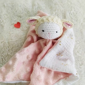 Op de afbeelding: Een handgemaakte babyknuffel met een gehaakt lammetjeshoofdje en een roze en witte pluche body. De deken heeft een zachte textuur en gouden details. Een klein rood hartje is ook zichtbaar.