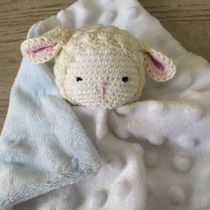 Op de afbeelding: Een zachte, witte babyknuffel met een gehaakt lammetjeshoofd. Het lam heeft roze oren en een roze neus. De deken is wit met verhoogde stippen en een lichtblauwe rand.