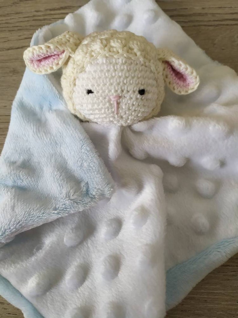 Op de afbeelding: Een zachte, witte babyknuffel met een gehaakt lammetjeshoofd. Het lam heeft roze oren en een roze neus. De deken is wit met verhoogde stippen en een lichtblauwe rand.