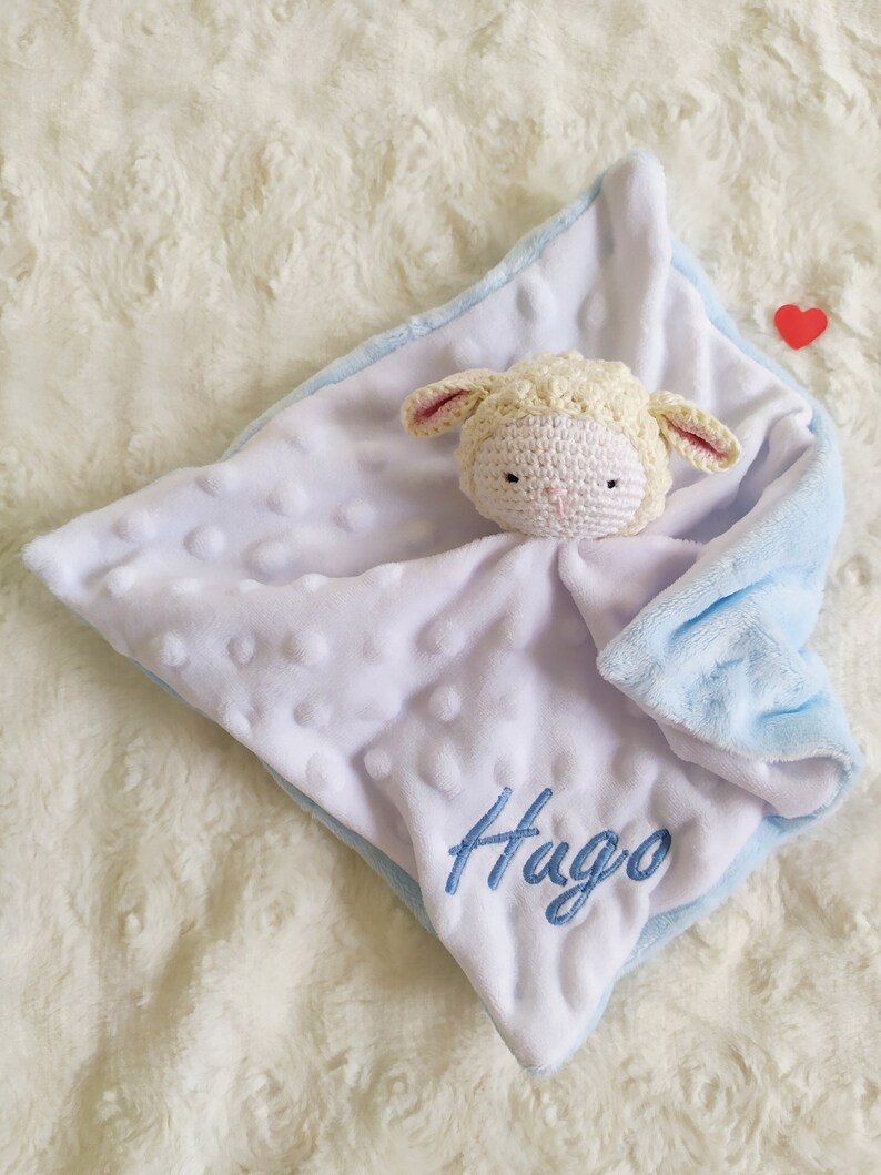 Op de afbeelding: Een witte en blauwe babydeken met een gehaakt lammetjeshoofdje. De deken heeft een zacht, getextureerd oppervlak, een blauwe rand en de naam "Hugo" geborduurd in blauw. Een klein rood hart is zichtbaar.