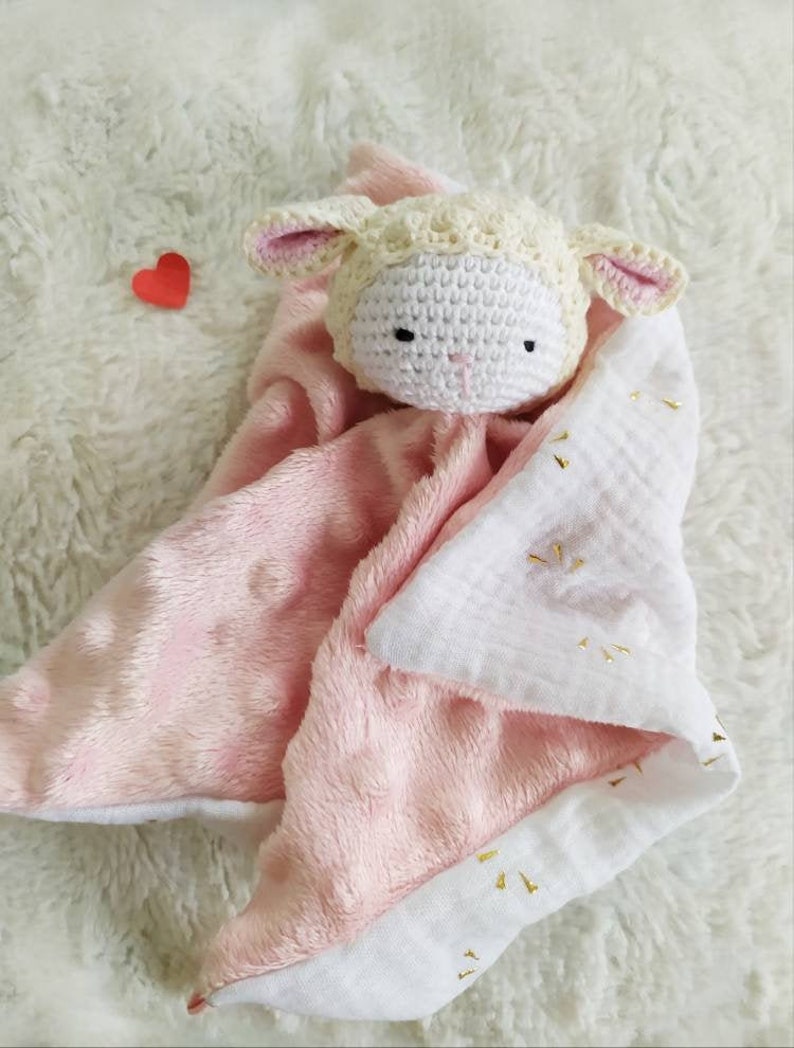 Op de afbeelding: Een handgemaakte babyknuffel met een gehaakt lammetjeshoofdje en een roze en witte pluche body. De deken heeft een zachte textuur en gouden details. Een klein rood hartje is ook zichtbaar.