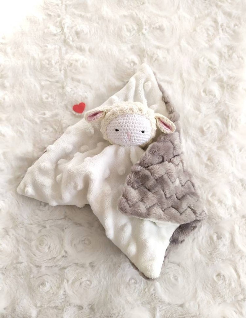 Op de afbeelding: Een witte en grijze babyknuffel met een gehaakt lammetjeshoofd. De deken heeft een zacht, getextureerd oppervlak met een grijs chevronpatroon op een hoek en een klein rood hart. Het lammetje heeft roze oren.