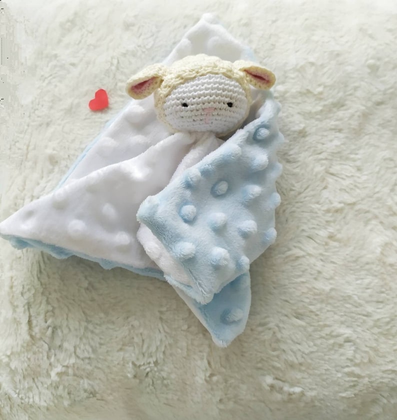 Op de afbeelding: Een babyknuffel met een gehaakt lammetjeshoofd. De deken is wit en lichtblauw, met een gestructureerd, gestippeld patroon op het blauwe gedeelte. Het lammetje heeft een cr&egrave;mekleurig hoofd en roze accenten op de oren.
