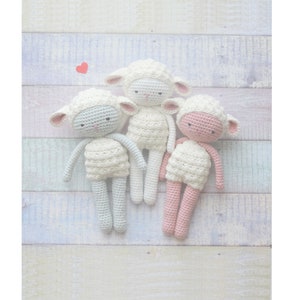 Op de afbeelding: Drie handgemaakte gehaakte schaapjes in wit, lichtblauw en roze. Elke pop heeft een witte body, een bijpassend hoofd en roze accenten op de oren en het gezicht. De poppen zijn gerangschikt op een lichtgekleurd, veelkleurig houten oppervlak.