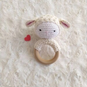 Op de afbeelding: Een handgemaakte gehaakte baby rammelaar in de vorm van een lammetje. Het lammetje is wit met roze oren en zwarte ogen. De rammelaar heeft een houten ring en een klein rood hart.