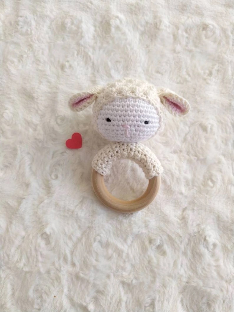 Op de afbeelding: Een handgemaakte gehaakte baby rammelaar in de vorm van een lammetje. Het lammetje is wit met roze oren en zwarte ogen. De rammelaar heeft een houten ring en een klein rood hart.