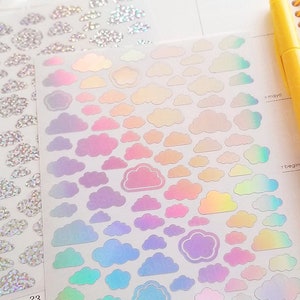 Pode incluir: Uma folha de adesivos de nuvens arco-íris iridescentes. Os adesivos estão dispostos em um degradê de azul claro a rosa. Os adesivos estão sobre um fundo branco. A folha está sobre um planejador com a palavra "maio" escrita nele.