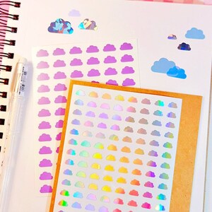 May include: A sheet of iridescent rainbow cloud stickers on a brown background. There are also sheets of purple and blue cloud stickers.