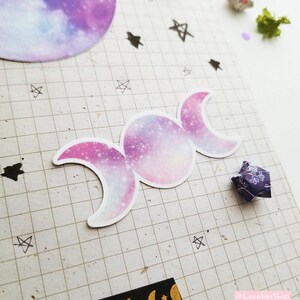 May include: A triple moon sticker with a galaxy print in shades of pink, purple, and blue. The sticker is on a gridded background.