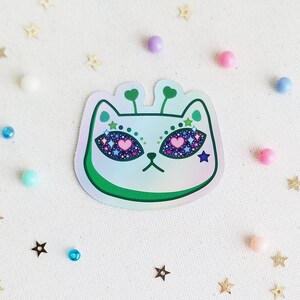 May include: A holographic sticker of a white cat with green antennas and sparkly eyes. The cat's eyes are filled with pink hearts and stars.