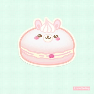 May include: A pink and white macaron with a cute cartoon llama face and a pink cherry on top. The macaron is decorated with white frosting and sprinkles.