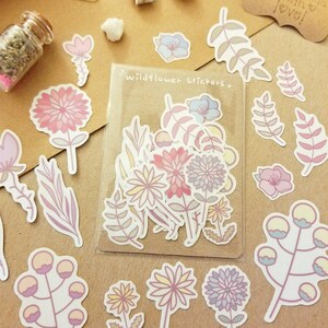 May include: A sheet of stickers featuring various pastel-colored wildflowers and foliage. The stickers are packaged in a clear plastic sleeve with the text "Wildflower Stickers" printed on it.