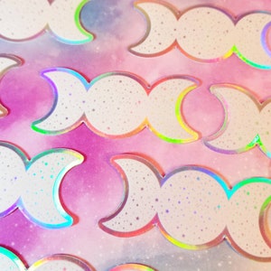 May include: A set of iridescent holographic stickers featuring the triple moon symbol. Each sticker has a white background with a glittery finish and a rainbow iridescent border.