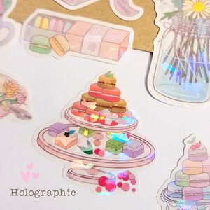 May include: A holographic sticker featuring a two-tiered cake stand with macarons, pastries, and cherries. The sticker has a pink and white background with a holographic effect.